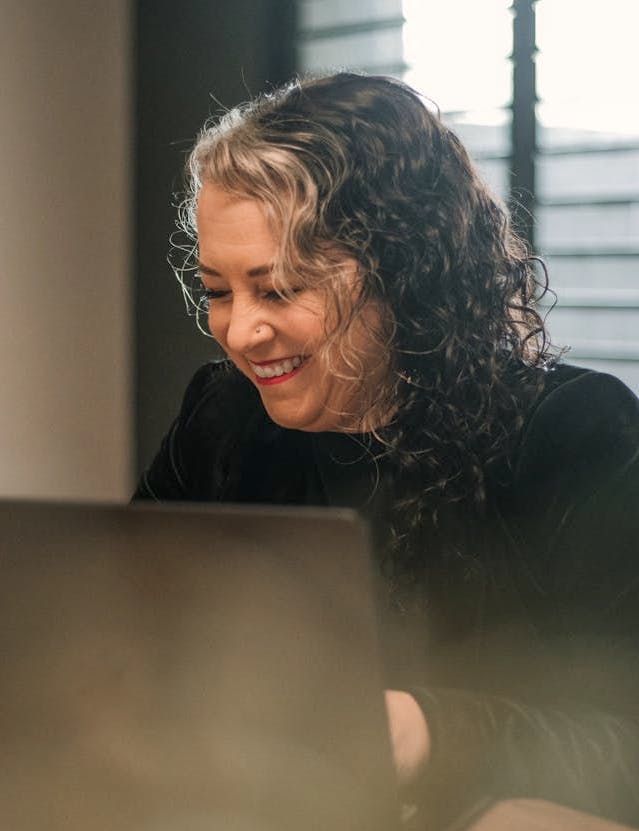 Close-up of a person sitting behind a laptop in an office space, grinning. Image by Prolific People on Pexels.