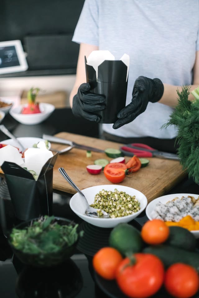 A person wearing black latex gloves presents a take-out box of food, over a table on which food is being prepared. Image by Mikhail Nilov on Pexels.